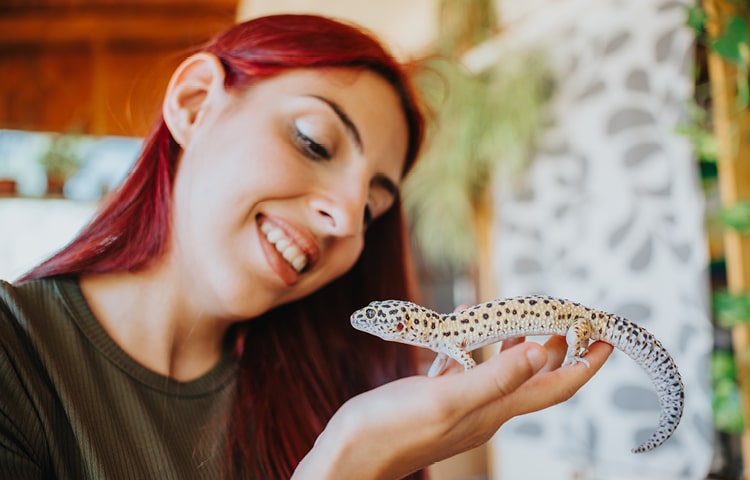 A young woman with red hair smiles as she holds a leopard gecko in her hand 